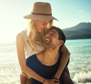 Lachendes Paar am Strand – sie trägt einen Strohhut, er trägt sie huckepack, im Hintergrund Meer und Berge.