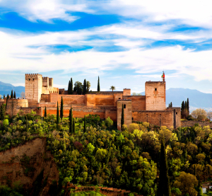 Die Alhambra in Granada, Spanien, umgeben von &uuml;ppigem Gr&uuml;n, mit den schneebedeckten Bergen der Sierra Nevada im Hintergrund unter blauem Himmel.