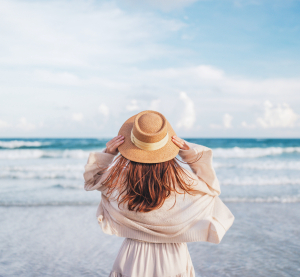 Frau mit Strohhut steht am Strand und blickt aufs Meer, während der Wind ihr Haar bewegt.