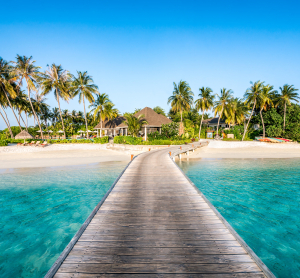 Steg f&uuml;hrt &uuml;ber t&uuml;rkisfarbenes Wasser zu einem tropischen Strandresort auf den Malediven, umgeben von Palmen und wei&szlig;em Sand bei strahlend blauem Himmel.