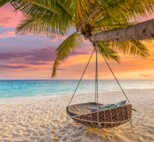 Hängesessel unter einer Palme an einem tropischen Sandstrand bei farbenfrohem Sonnenuntergang.