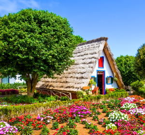Traditionelles Strohdachhaus zwischen bunten Blumenbeeten und grünen Bäumen.