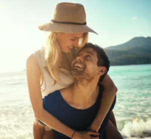Lachendes Paar am Strand – sie trägt einen Strohhut, er trägt sie huckepack, im Hintergrund Meer und Berge.