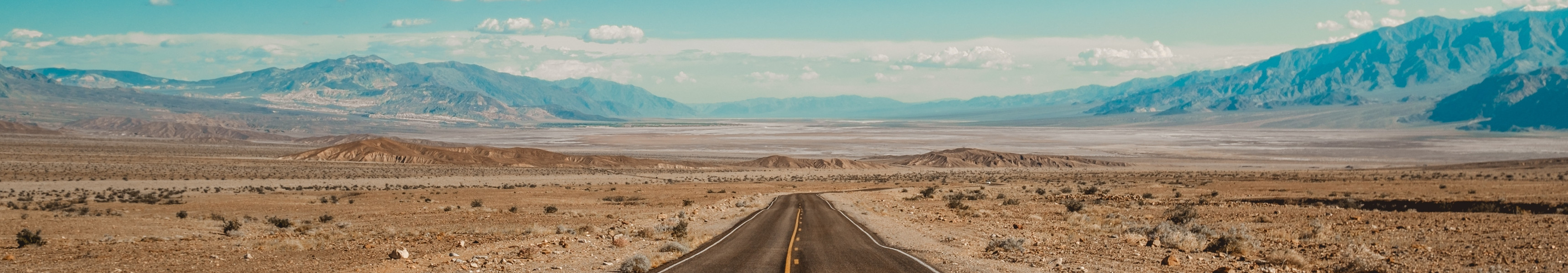 Eine endlos wirkende Straße führt durch die karge Wüstenlandschaft des Death Valley in Kalifornien, USA, mit Bergen am Horizont unter einem klaren blauen Himmel.