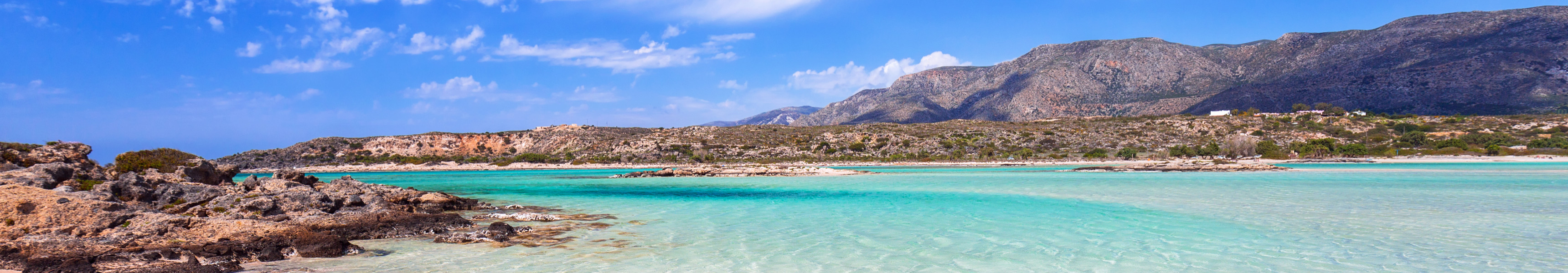 Traumhafte Lagune von Elafonissi auf Kreta mit rosa schimmerndem Sand, türkisblauem Wasser und felsiger Küste vor einer Bergkulisse unter blauem Himmel mit weißen Wolken.