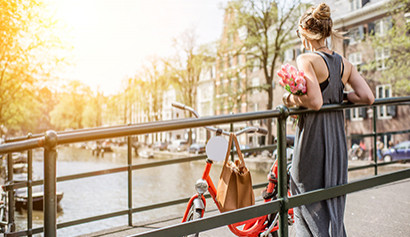 Frau mit Blumenstrauß lehnt an einem Brückengeländer in Amsterdam, neben ihr ein rotes Fahrrad.