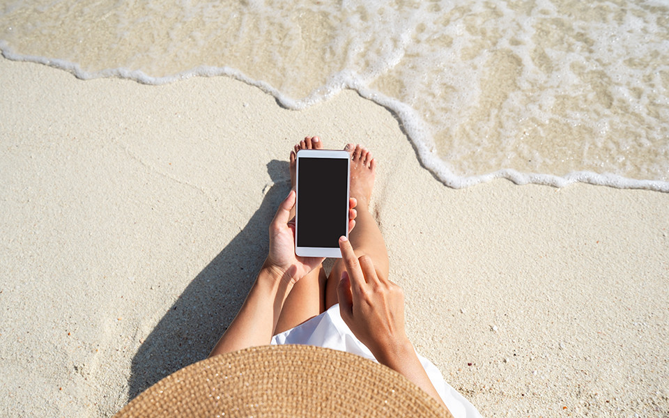 Frau sitzt mit einem Smartphone in der Hand am Sandstrand direkt am Wasser, ihre F&uuml;&szlig;e ber&uuml;hren den nassen Sand