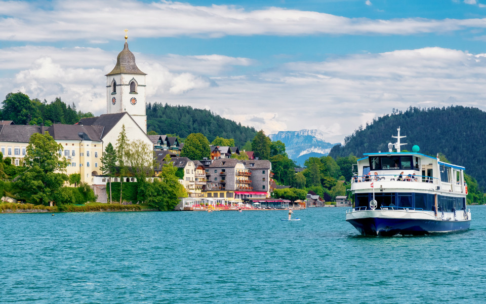 Der Wolfgangsee mit einem Ausflugsboot im Vordergrund und einer malerischen Landschaft, einschlie&szlig;lich der markanten Kirche und der Berge im Hintergrund.