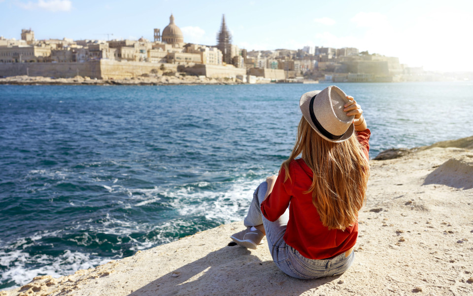 Sch&ouml;ne junge Frau mit Hut sitzt auf einem Stein am Meer und blickt auf einen atemberaubenden Panoramablick auf die Stadt Valletta auf Malta