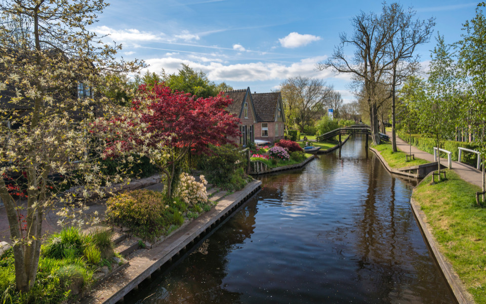 Kleines Backsteinhaus mit Garten an einem ruhigen Kanal in Giethoorn, umgeben von blühenden Bäumen und Fußgängerbrücken.